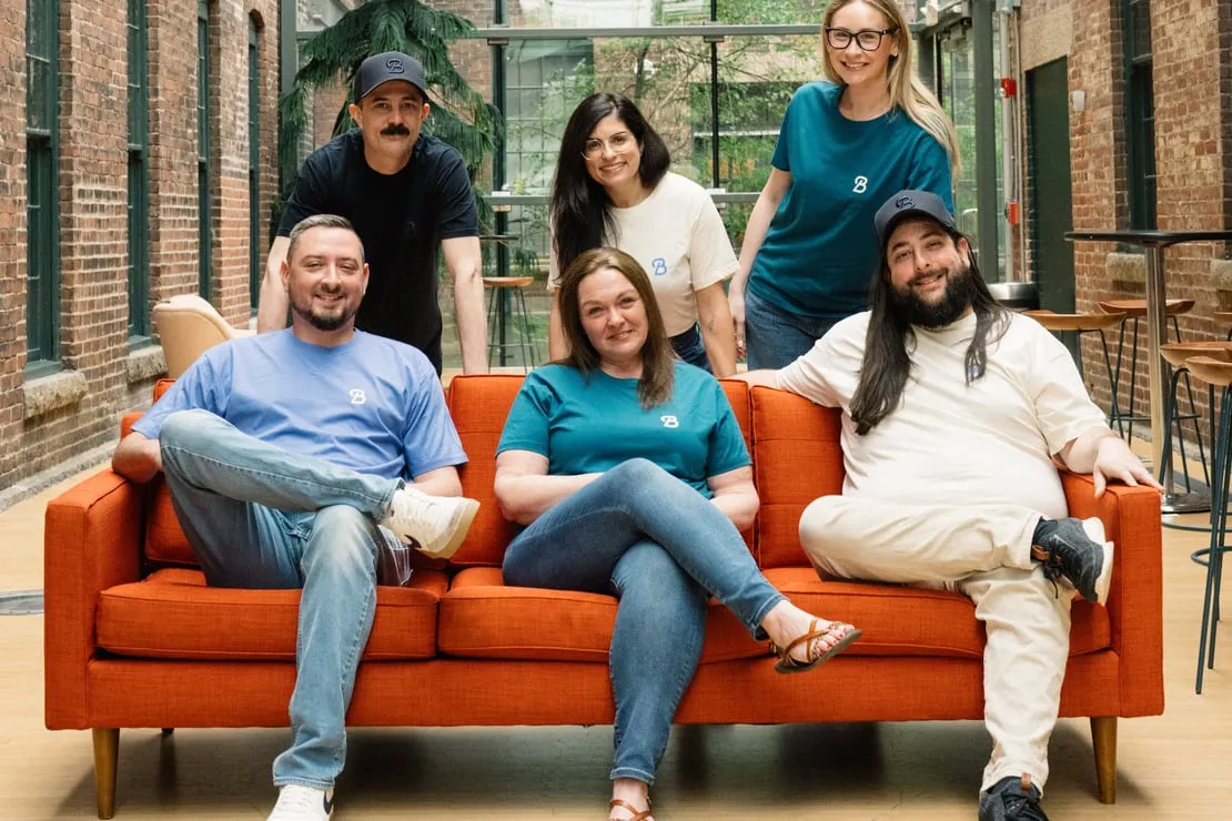 Beeline Equity Now team — three seated on an orange couch with three colleagues standing behind, in a brick-and-glass atrium
